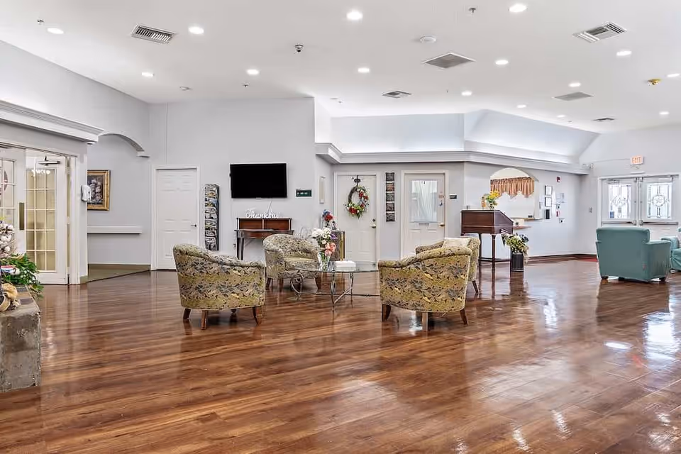 Spacious bright communal lounge with floral armchairs arranged around glass tables on polished wood floors, a wall-mounted TV and a piano.