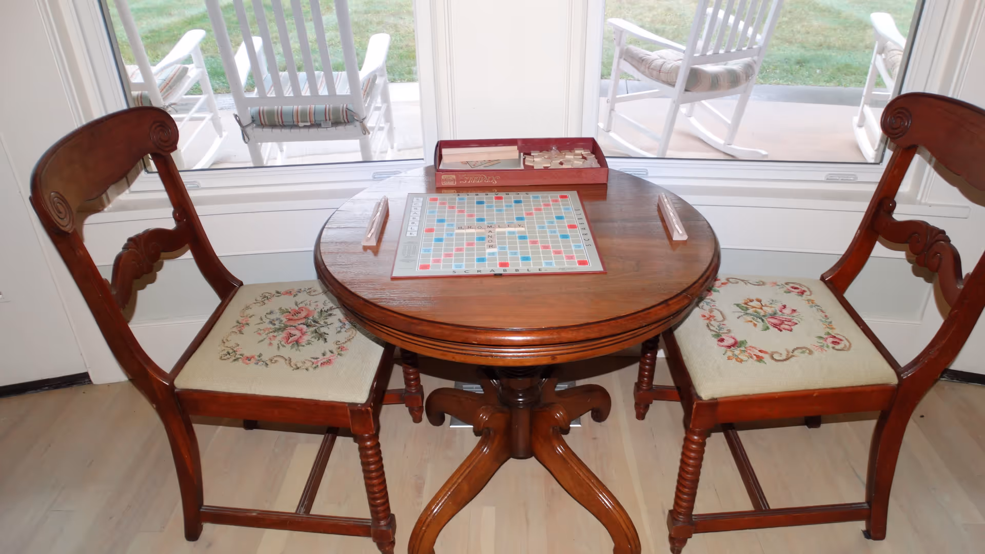 A small round wooden table with a Scrabble board game set up on it, flanked by two wooden chairs with floral upholstered seats. Behind the table is a large window showing a patio with white rocking chairs and green grass outside.