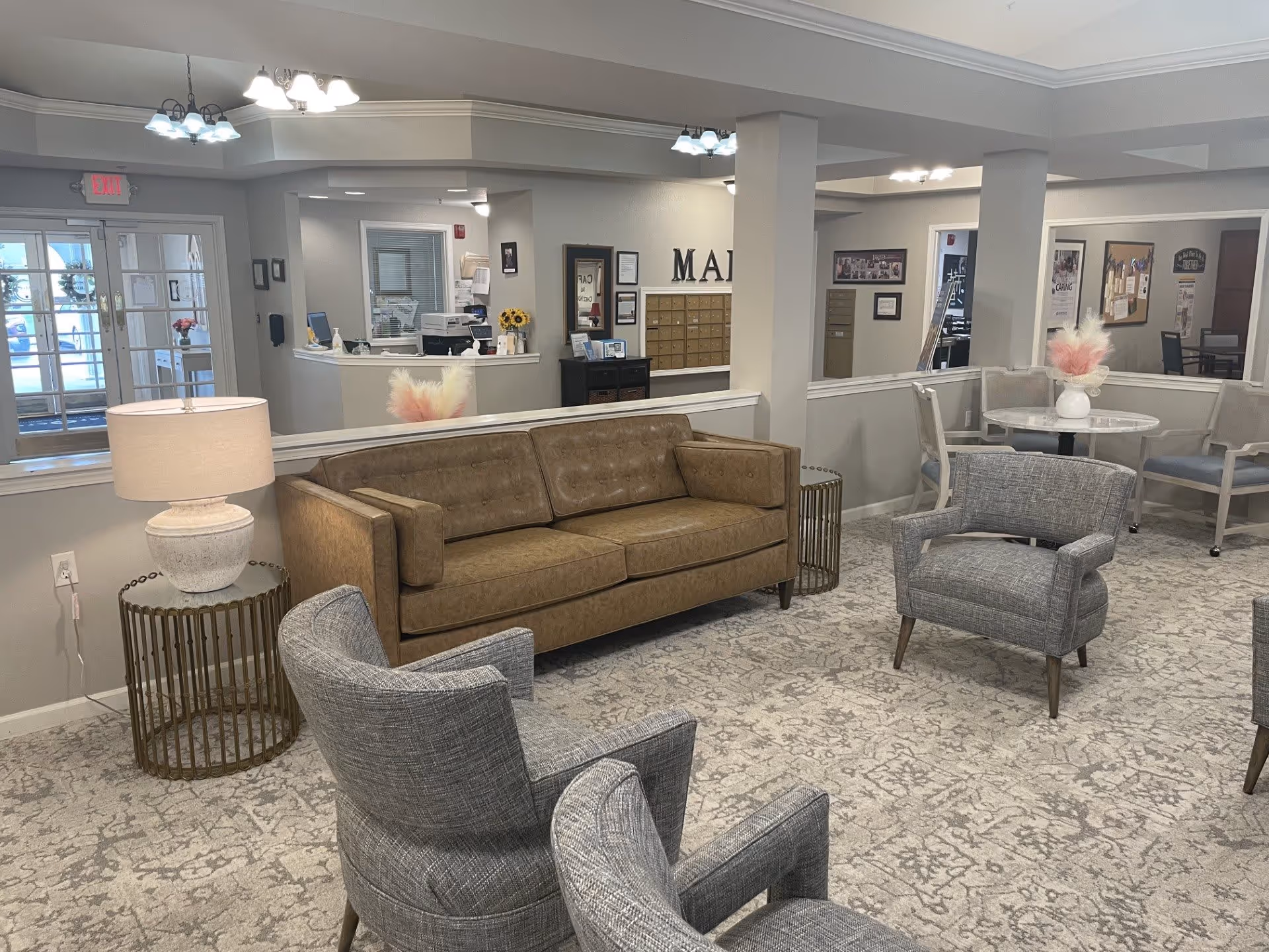 A cozy senior living facility common area with a brown leather sofa, three gray upholstered armchairs, a round side table with a lamp, and a small round dining table with chairs. The background shows a reception desk, mailboxes, and an entrance door with glass panels.