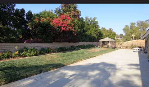 Outdoor area with a concrete patio, green grass lawn, and a garden bed with various plants and flowers along a brick wall. There is a gazebo with seating in the background and trees surrounding the area under a clear blue sky.