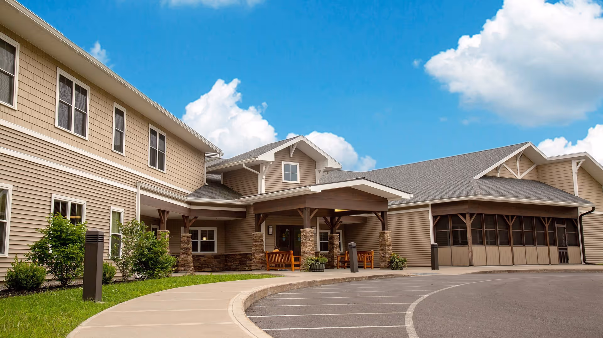 Front entrance of a residential care building with a covered porch, outdoor seating, and landscaped grounds under a blue sky.