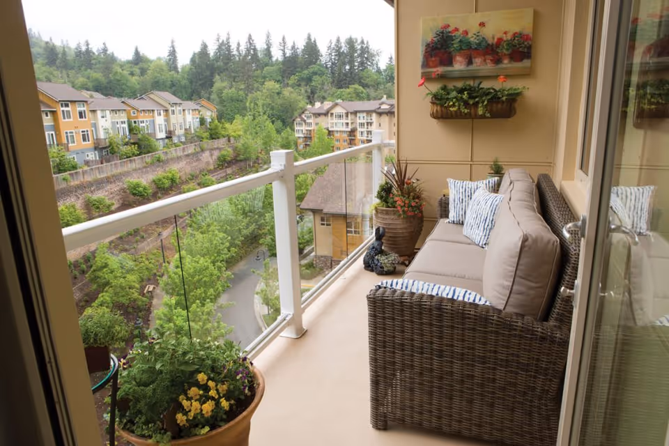 A furnished balcony with a wicker sofa, potted plants, and a glass railing overlooking neighboring buildings and trees.
