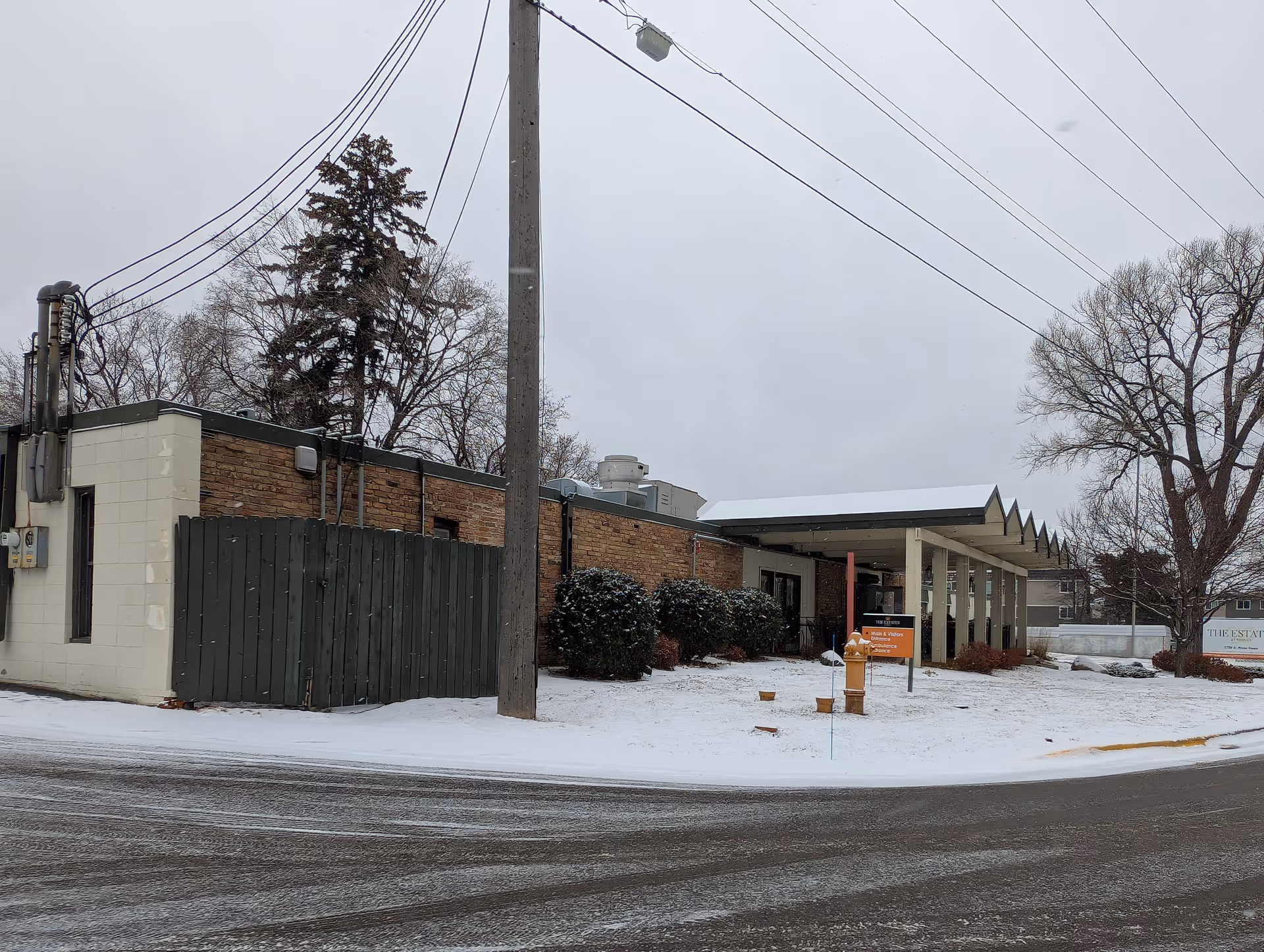 Exterior view of The Estates at Fridley building in winter showing a snow-covered lawn, covered entrance canopy, and surrounding trees.