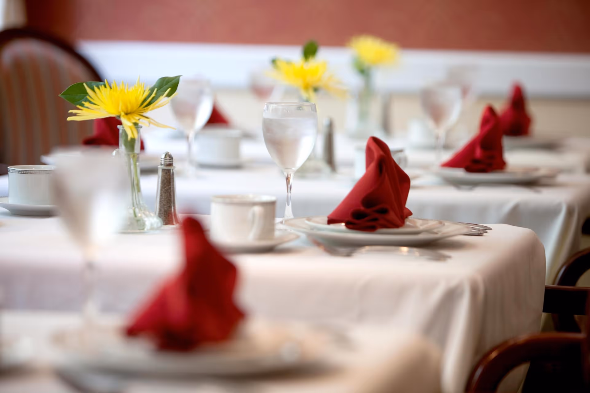 Tables in a dining room set with white tablecloths, red folded napkins, water glasses and yellow flower centerpieces.