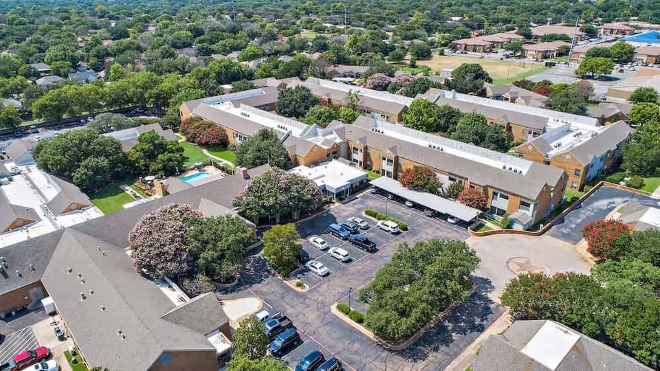 Aerial view of Morada Temple senior living facility showing multiple connected buildings with brown roofs, surrounded by trees and greenery. There is a parking lot with several cars, a swimming pool, and well-maintained landscaping.