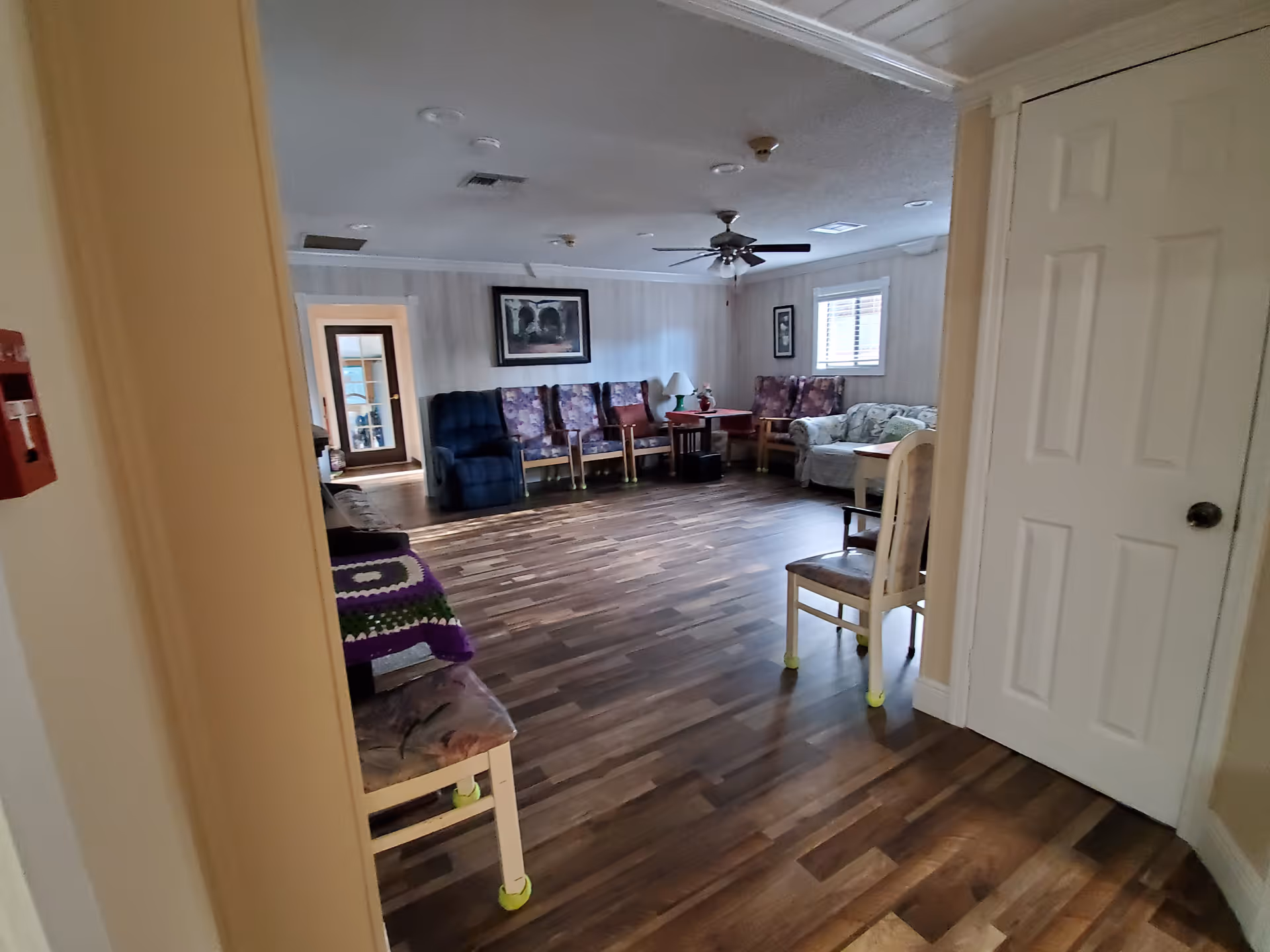 Wide view of a common sitting room with sofas and chairs around the perimeter, wood-look flooring and a ceiling fan.