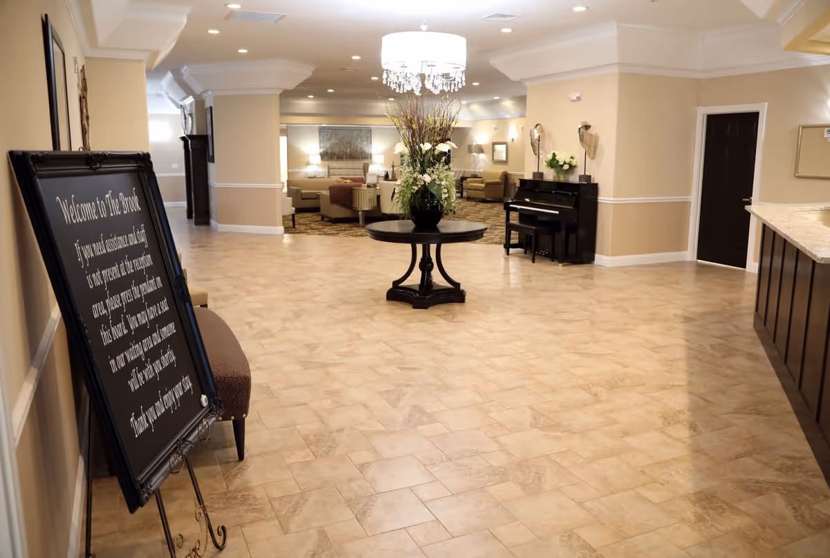 Spacious and well-lit interior common area of The Brook of Rogers City featuring a round table with a large floral arrangement in the center, a chandelier overhead, a black upright piano against the wall, and comfortable seating areas with sofas and chairs in the background. A large framed welcome sign is displayed on an easel near the entrance.