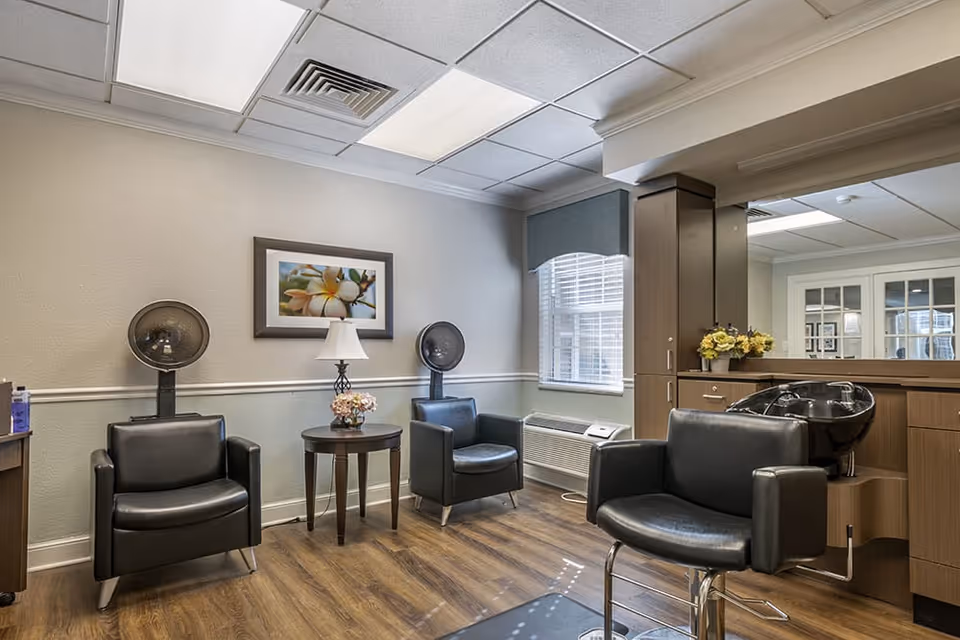 Interior of a hair salon area in a senior living facility featuring two black leather chairs with hair dryers behind them, a small round table with a lamp and flowers, a window with blinds and a blue valance, and a hair washing station with a black chair and sink. The room has wood flooring, light-colored walls, and a framed flower picture on the wall.