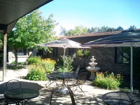 Outdoor courtyard with tables, chairs, umbrellas, and a tiered fountain near a brick building.
