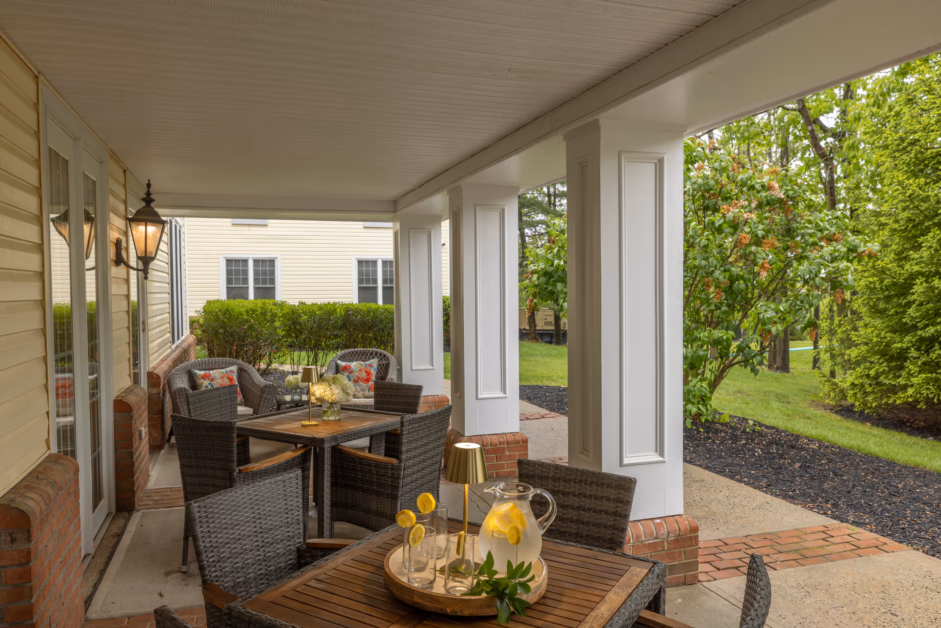 Covered outdoor patio with wicker dining tables and chairs, decorative columns, and a pitcher of lemon water overlooking landscaped grounds.