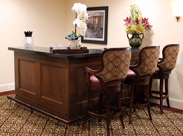 A wooden bar counter with a dark granite top, accompanied by three upholstered bar stools with patterned backs and seats. On the counter are decorative items including a white orchid plant, a green vase with flowers, and a glass container with black utensils. The room has patterned carpet flooring and a framed picture on the wall behind the bar.