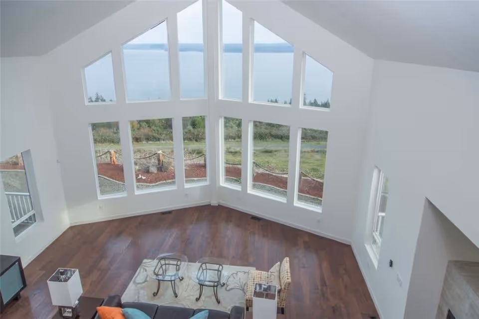 Spacious living room with large floor-to-ceiling windows offering a scenic view of greenery and water outside. The room has wooden flooring, a patterned area rug, a sofa with colorful cushions, two glass-top side tables, and a patterned armchair.