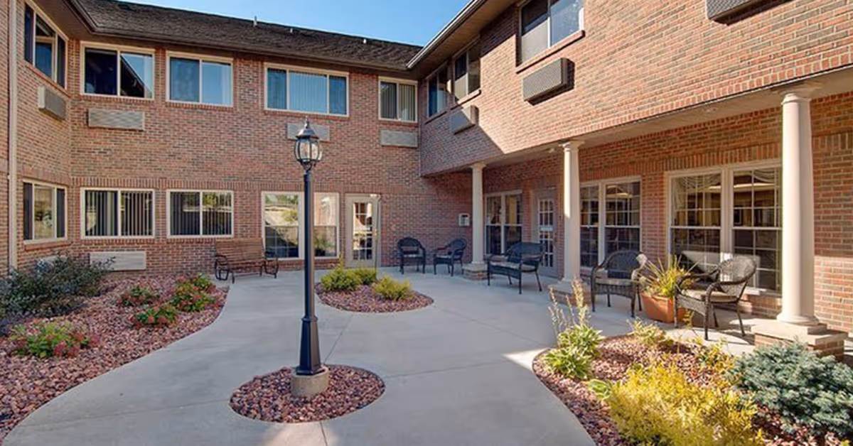 Sunlit brick courtyard with a central lamppost, outdoor seating, and landscaped planters at a senior living facility.