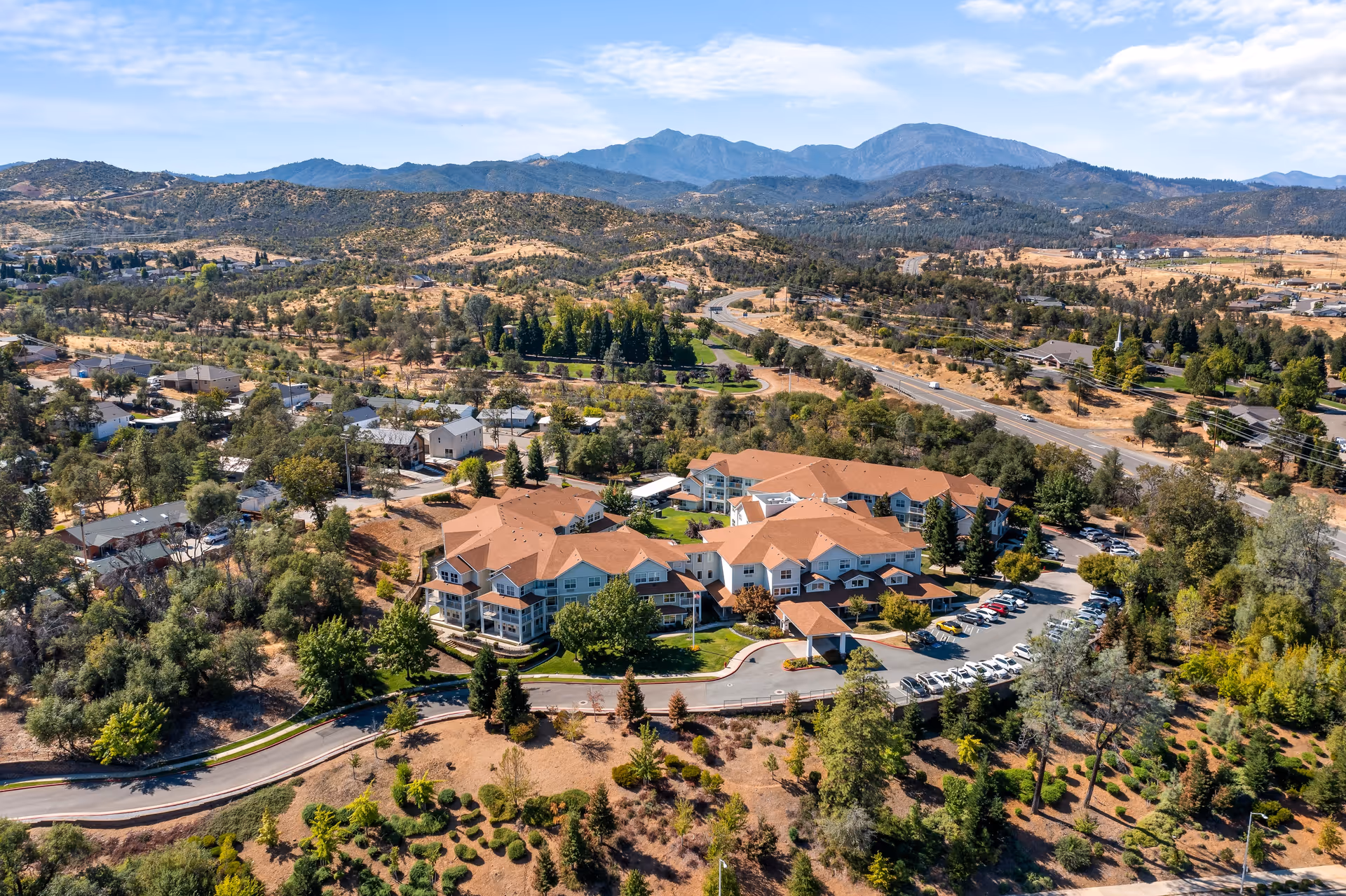 Aerial view of Shasta Estates Senior Living facility surrounded by trees and hills, with multiple buildings featuring red roofs, parking lots with cars, and a winding road leading to the entrance. The background shows mountainous terrain under a partly cloudy sky.
