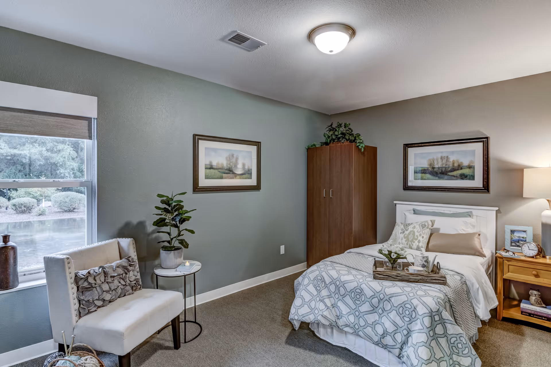 A cozy furnished bedroom with a bed, bedside table and lamp, an armchair by a window, and framed artwork on blue-gray walls.
