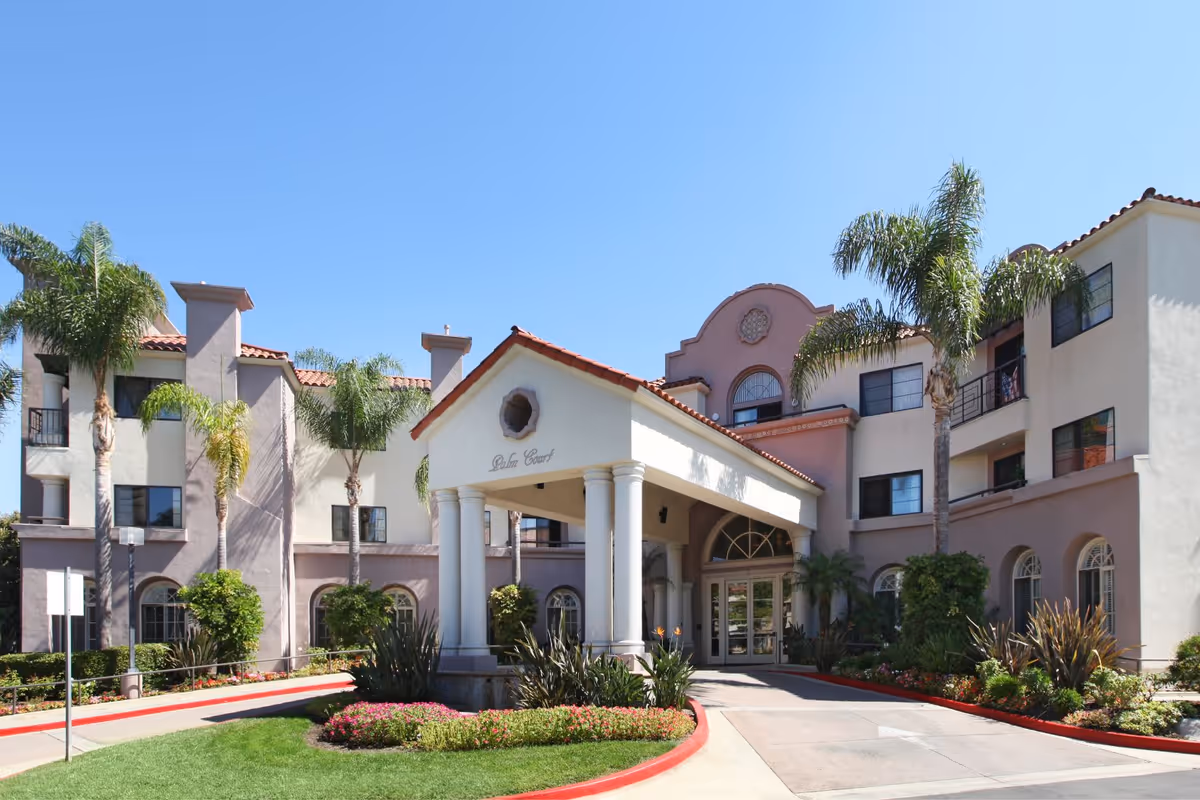 Front entrance of a Mediterranean-style senior living building named Palm Court with a porte-cochère, palm trees, and landscaped driveway.