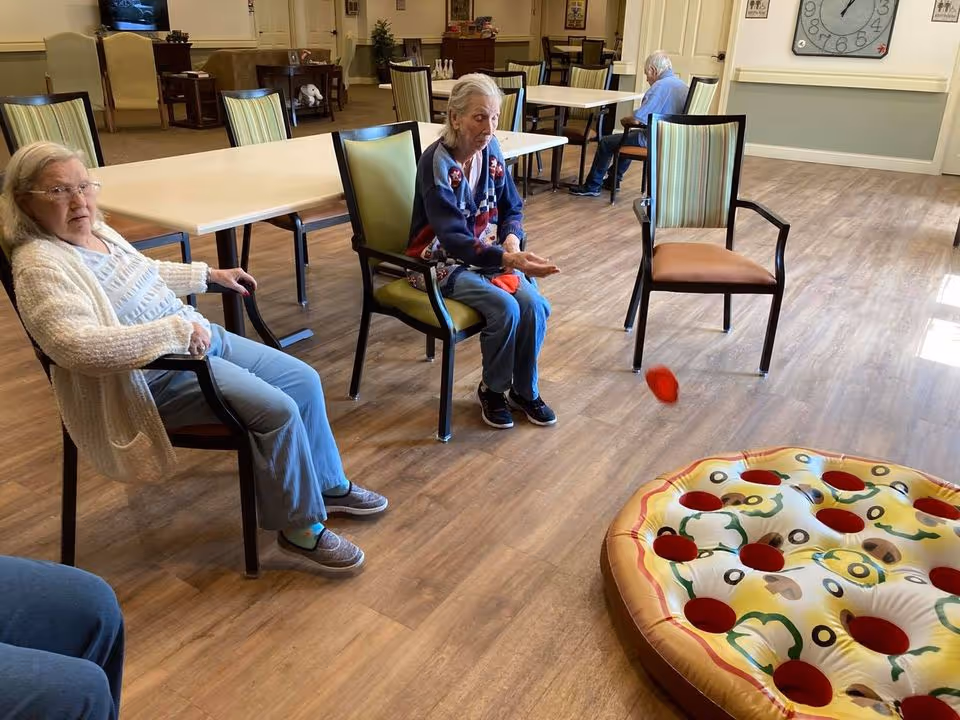 Three elderly individuals seated in a common room with wooden flooring and tables. Two women are sitting on chairs in the foreground, one tossing a red bean bag towards an inflatable pizza-shaped target on the floor. Another person is seated at a table in the background.