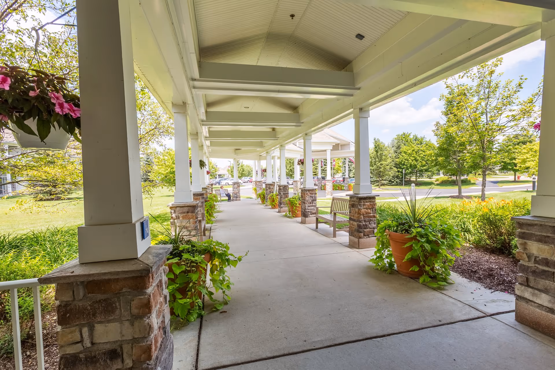 Covered outdoor walkway with white pillars and stone bases, lined with potted plants and benches, overlooking a green landscaped area with trees and a clear sky.