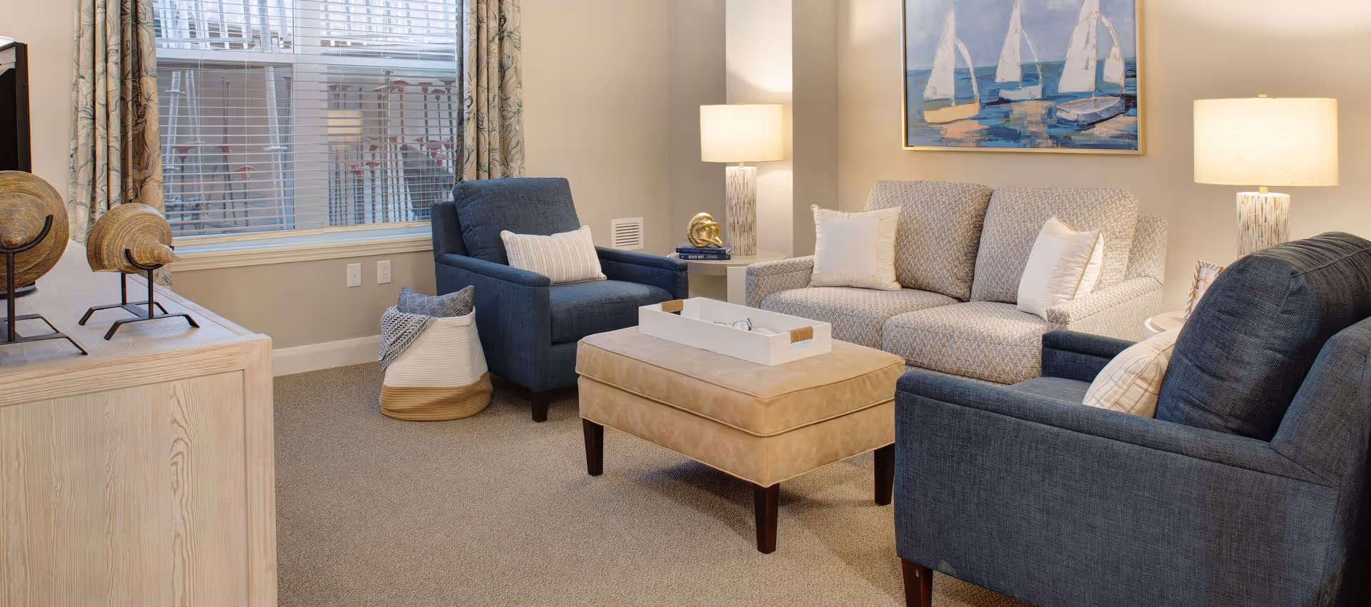 Cozy living room with a sofa, two blue armchairs, an ottoman coffee table, lamps, and a window.