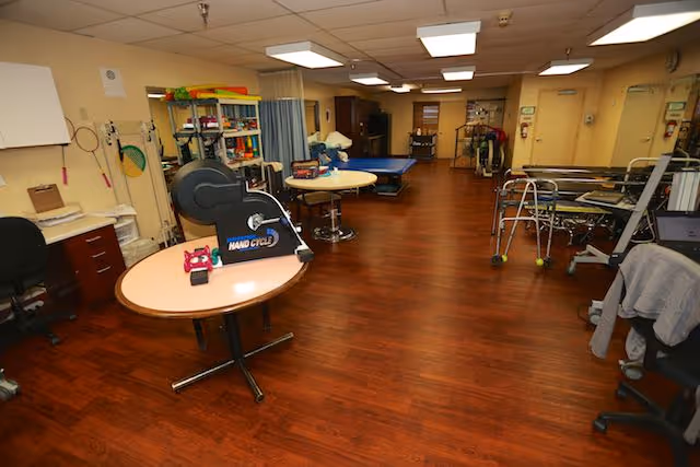 A spacious room with wooden flooring containing various physical therapy and rehabilitation equipment including a hand cycle on a round table, walkers, exercise balls, and therapy mats. The room is well-lit with ceiling lights and has a desk and chairs along the walls.