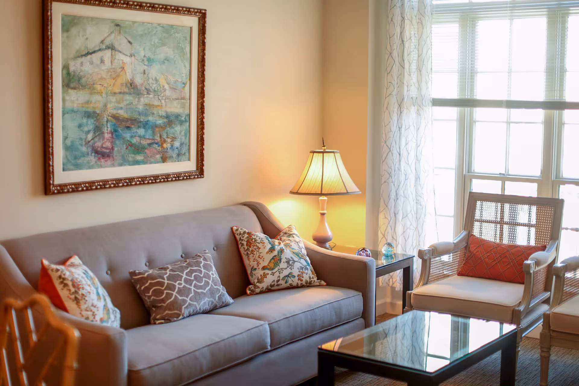 A cozy living room with a gray sofa adorned with patterned throw pillows, a wooden armchair with a red cushion, a glass coffee table, a side table with a lamp, and a large window with sheer curtains letting in natural light. A framed painting hangs on the wall above the sofa.