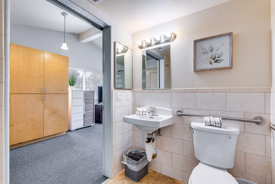 A clean and accessible bathroom with a white toilet and wall-mounted sink. The walls are tiled halfway up, and there is a grab bar next to the toilet. Above the sink is a mirror with lights, and a framed botanical picture hangs on the wall. The bathroom opens into a carpeted room with wooden cabinets and a window with blinds.