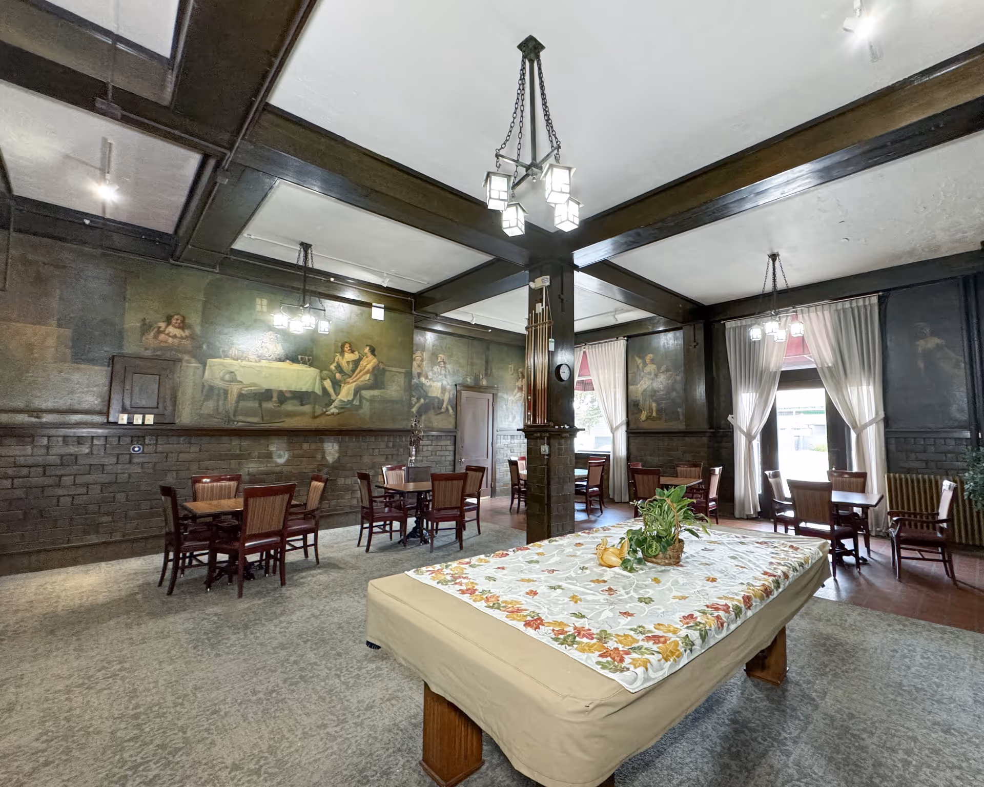 A spacious room with a covered pool table in the center, decorated with a floral tablecloth and a small plant. Surrounding the pool table are several wooden tables and chairs. The walls feature large murals depicting people in historical attire, and there are large windows with white curtains letting in natural light. The ceiling has exposed dark wooden beams and hanging light fixtures.