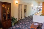 Interior hallway of a senior living facility with floral carpet, cream-colored walls, and several doors along the corridor. A wooden cabinet with glass doors and decorative items inside is on the left, next to a potted plant. On the right side, there is a railing overlooking a lower level and a small wooden table with magazines on it.