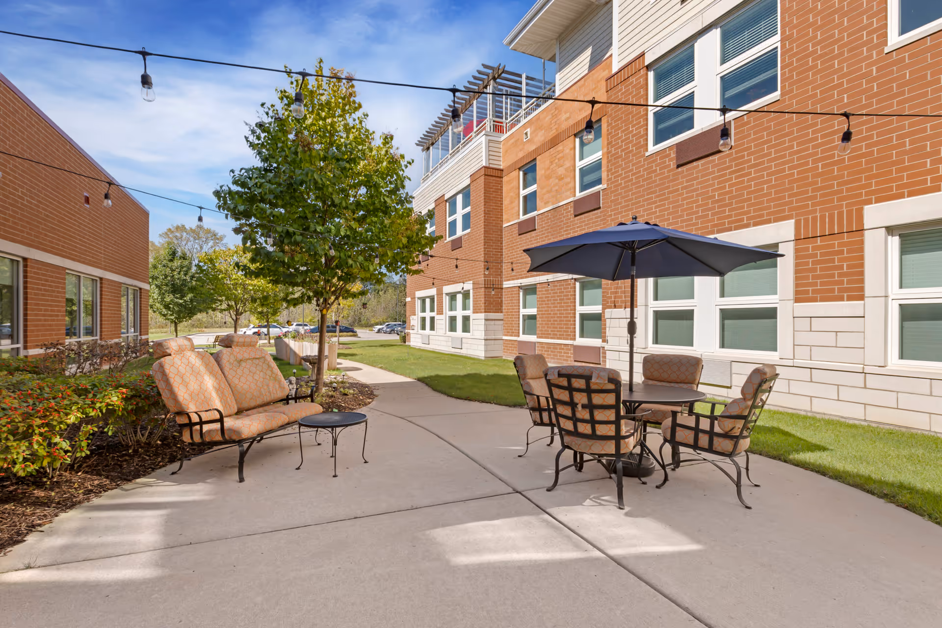 Outdoor patio area with cushioned seating including a loveseat and chairs around a table with a large umbrella. The patio is adjacent to a brick building with multiple windows and string lights hanging overhead. There are trees and shrubs along the walkway.