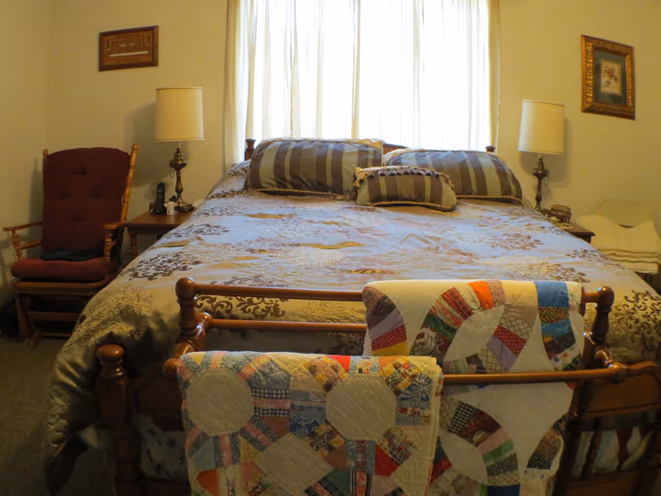 A cozy bedroom with a large wooden bed covered in patterned bedding and quilts draped over the footboard, flanked by bedside lamps and a rocking chair.