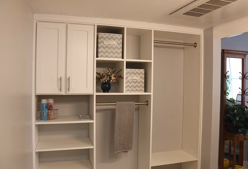 A white built-in closet organizer with shelves, cabinets, and hanging rods. Two gray and white chevron-patterned storage bins and a small blue vase with flowers are placed on the shelves. A gray towel hangs on one of the rods. To the right, a wooden framed mirror and some green plants are partially visible.