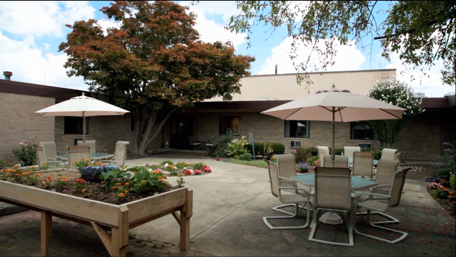 Outdoor patio area with tables and chairs under large umbrellas, surrounded by flower beds and greenery, with a building in the background and a partly cloudy sky.