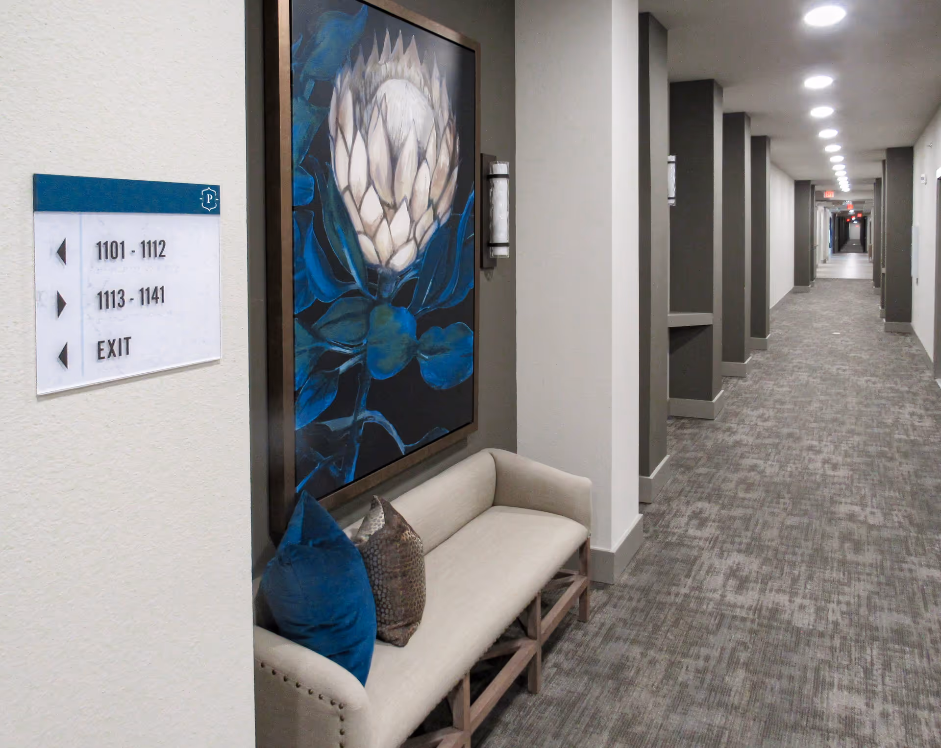 A long, well-lit hallway in a senior living facility with a beige carpet and gray pillars along the right side. On the left side, there is a beige bench with two decorative pillows beneath a large framed painting of a flower with blue leaves. A sign on the wall indicates room numbers 1101-1112 and 1113-1141, as well as the exit direction.
