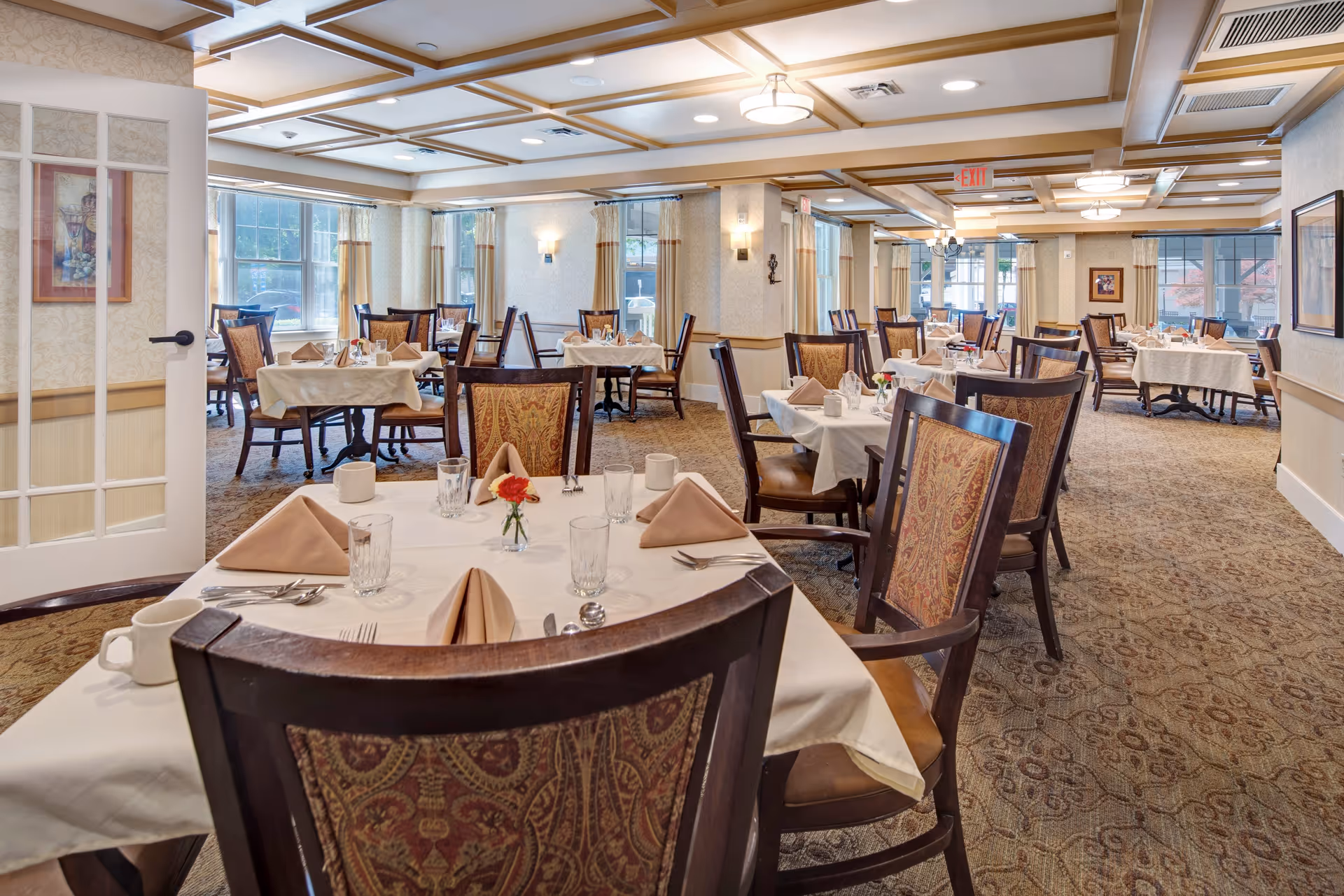 A spacious dining room in a senior living facility with multiple tables covered in white tablecloths, each set with beige folded napkins, glassware, silverware, and small flower vases. The room has patterned carpet, large windows allowing natural light, and wooden chairs with ornate upholstery. The ceiling features wooden beams and recessed lighting.