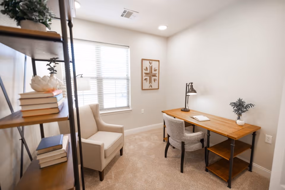A small, cozy room with beige carpet and white walls featuring a wooden desk with a lamp, an open book, and a small plant. There is a comfortable upholstered chair at the desk and another armchair near a window with blinds. A wooden shelf with books and decorative items is visible on the left side of the image, and a framed geometric artwork hangs on the wall.