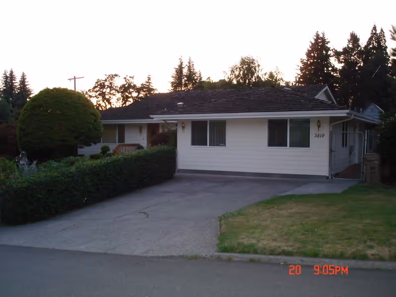 Exterior view of a single-story residential building with white siding and a dark shingled roof, surrounded by trees and bushes. There is a driveway leading up to the house and a small lawn area on the right side. The house number 3819 is visible near the front door.