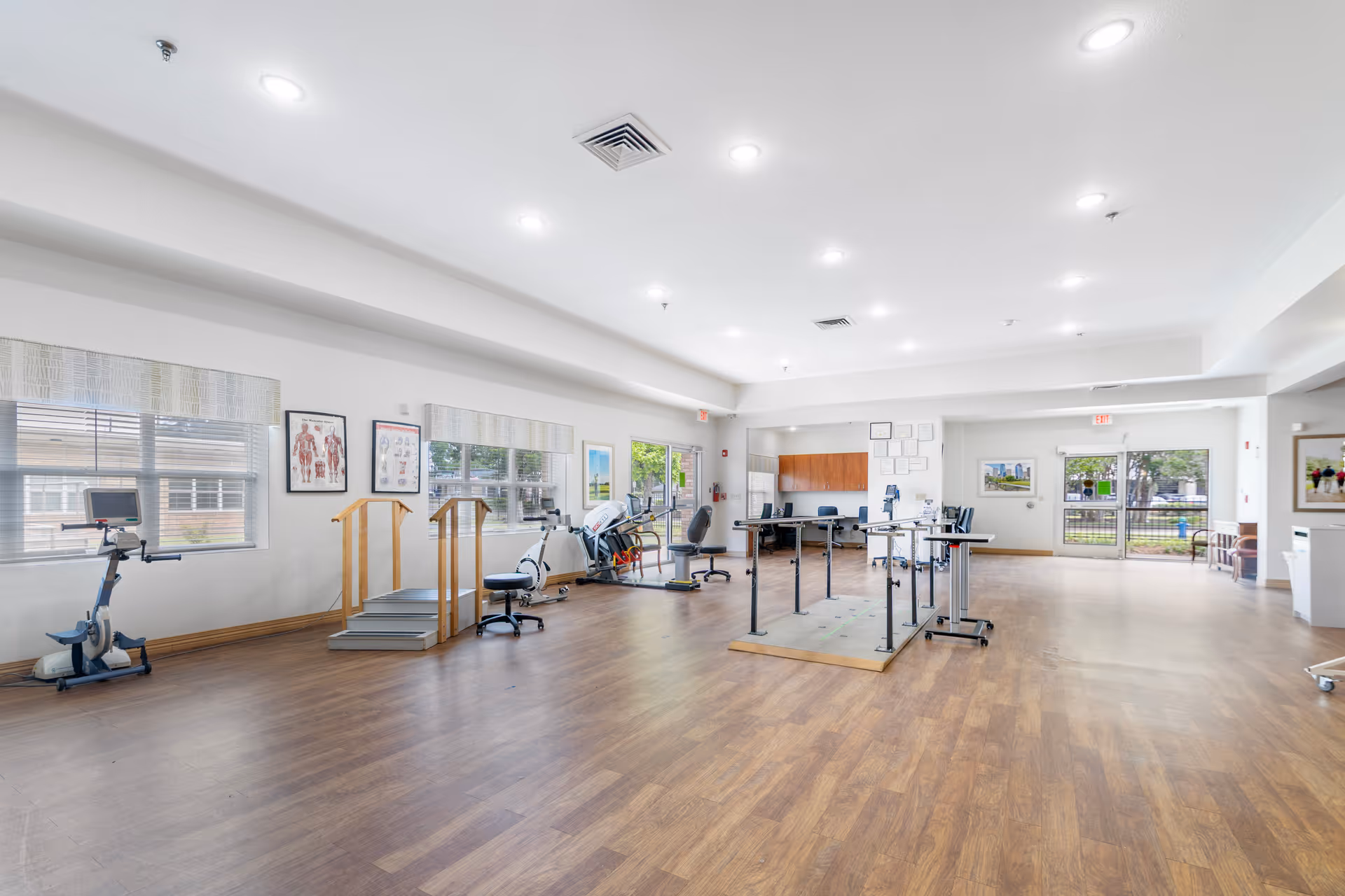 Spacious rehabilitation therapy room with exercise equipment, parallel bars, and large windows.