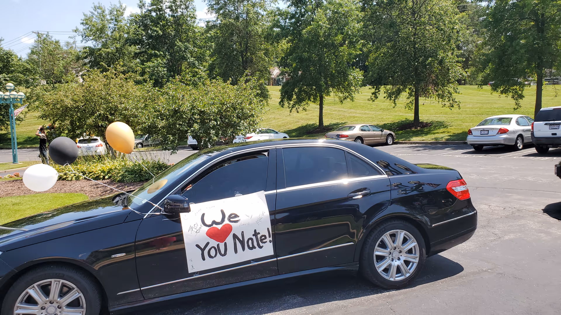 A black sedan parked in a parking lot with a handmade sign taped to the driver's side door that reads 'We ♥ You Nate!'. There are black, white, and gold balloons tied to the car. Trees and other parked cars are visible in the background under a clear sky.
