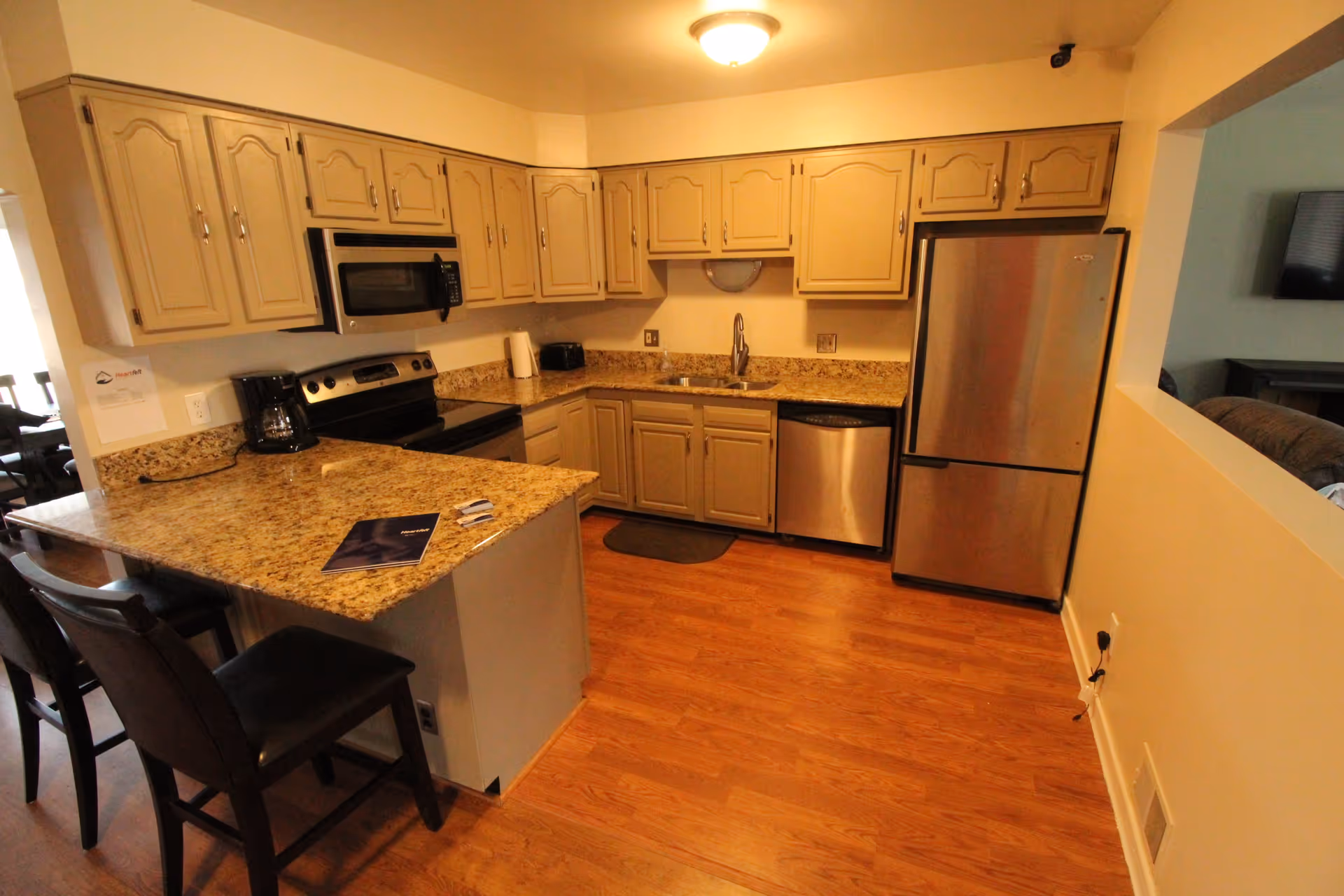 A kitchen with beige cabinets, granite countertops, and wooden flooring. The kitchen features a stainless steel refrigerator, dishwasher, microwave, and stove. There are two black chairs at the counter, a coffee maker, and a small window opening to an adjacent room with a TV and couch visible.