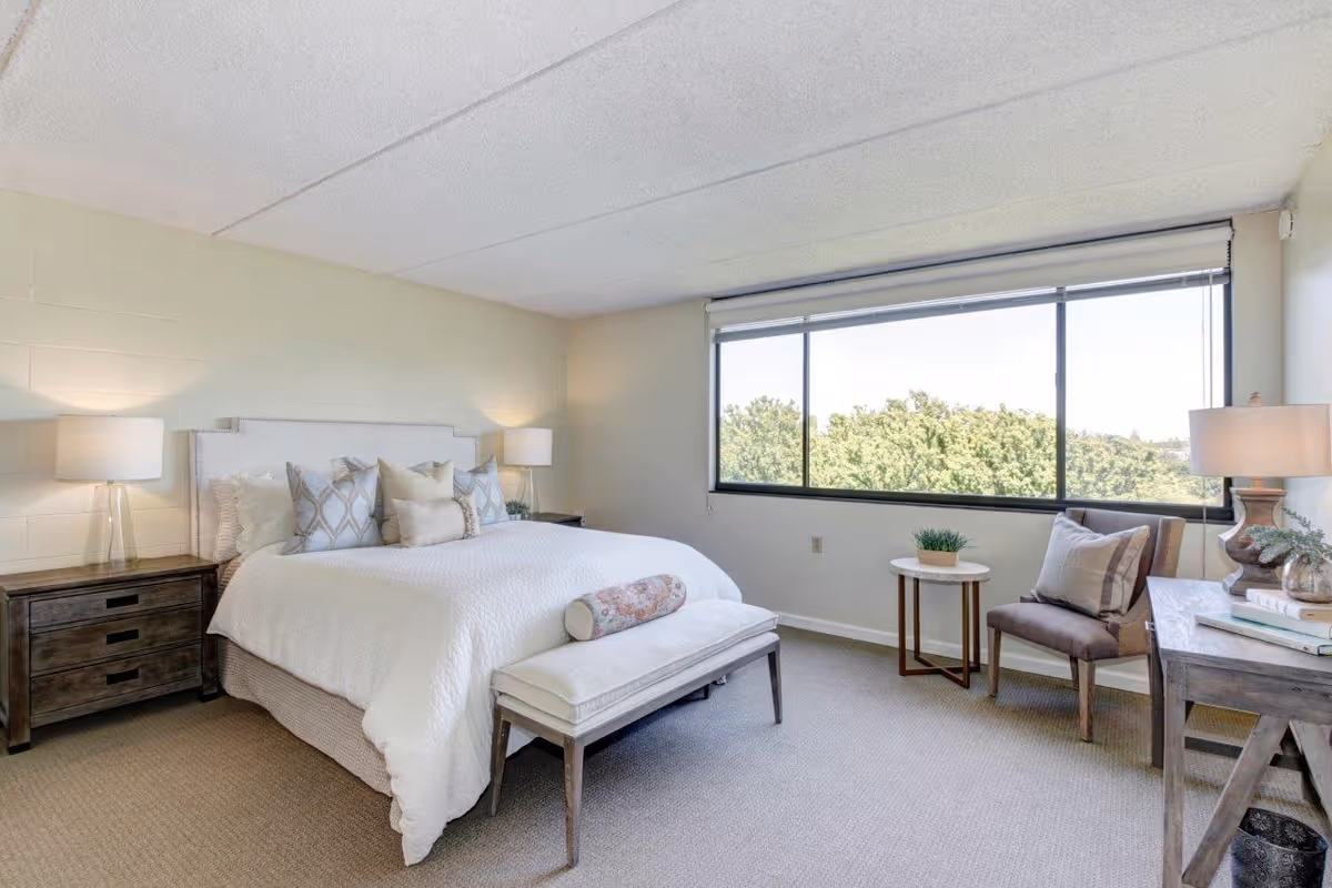 A bright and cozy bedroom with a large window showing green trees outside. The room features a neatly made bed with white bedding and multiple decorative pillows, flanked by two wooden nightstands each with a lamp. At the foot of the bed is a cushioned bench. There is a small round table with a plant and a cushioned chair next to the window. A wooden desk with a lamp, books, and decorative items is positioned on the right side of the room.