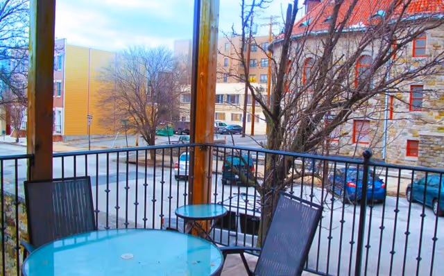 View from a balcony with a round glass table and two chairs, overlooking a parking lot with several parked cars and buildings in the background. Leafless trees are visible near the balcony railing.