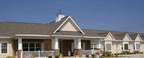 Exterior view of a single-story assisted living facility building with a covered entrance, white columns, and a small cupola on the roof under a clear blue sky.
