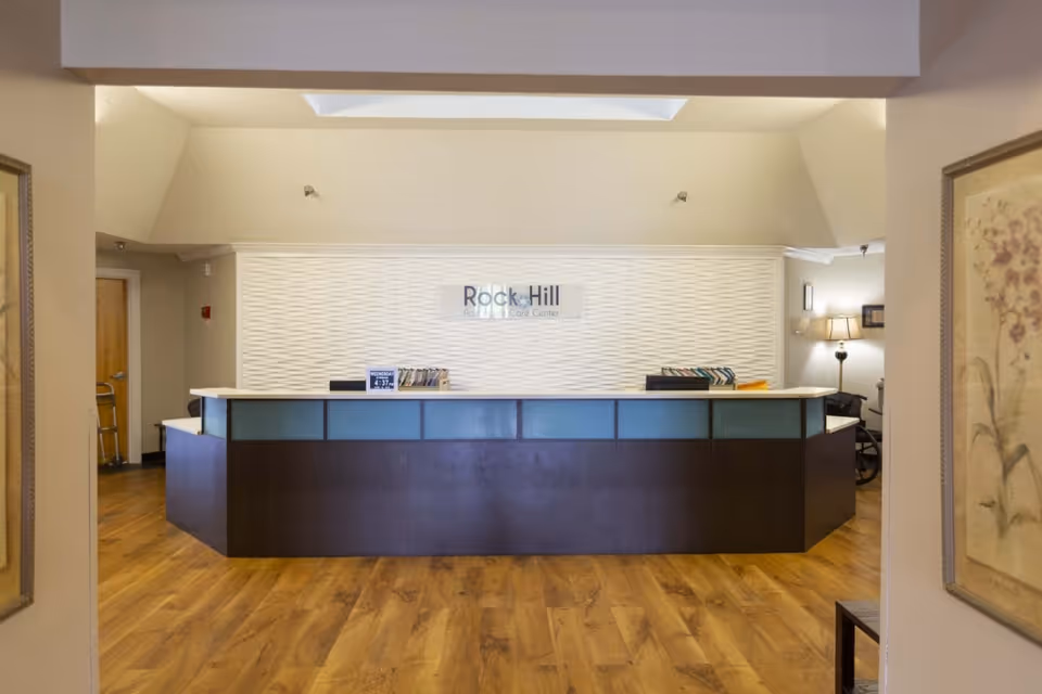 Reception desk area inside Rock Hill Post Acute Care Center with a modern dark wood and frosted glass counter, light wood flooring, and a textured white wall behind the desk displaying the facility's name. The space is well-lit with a skylight above and framed artwork on the walls.