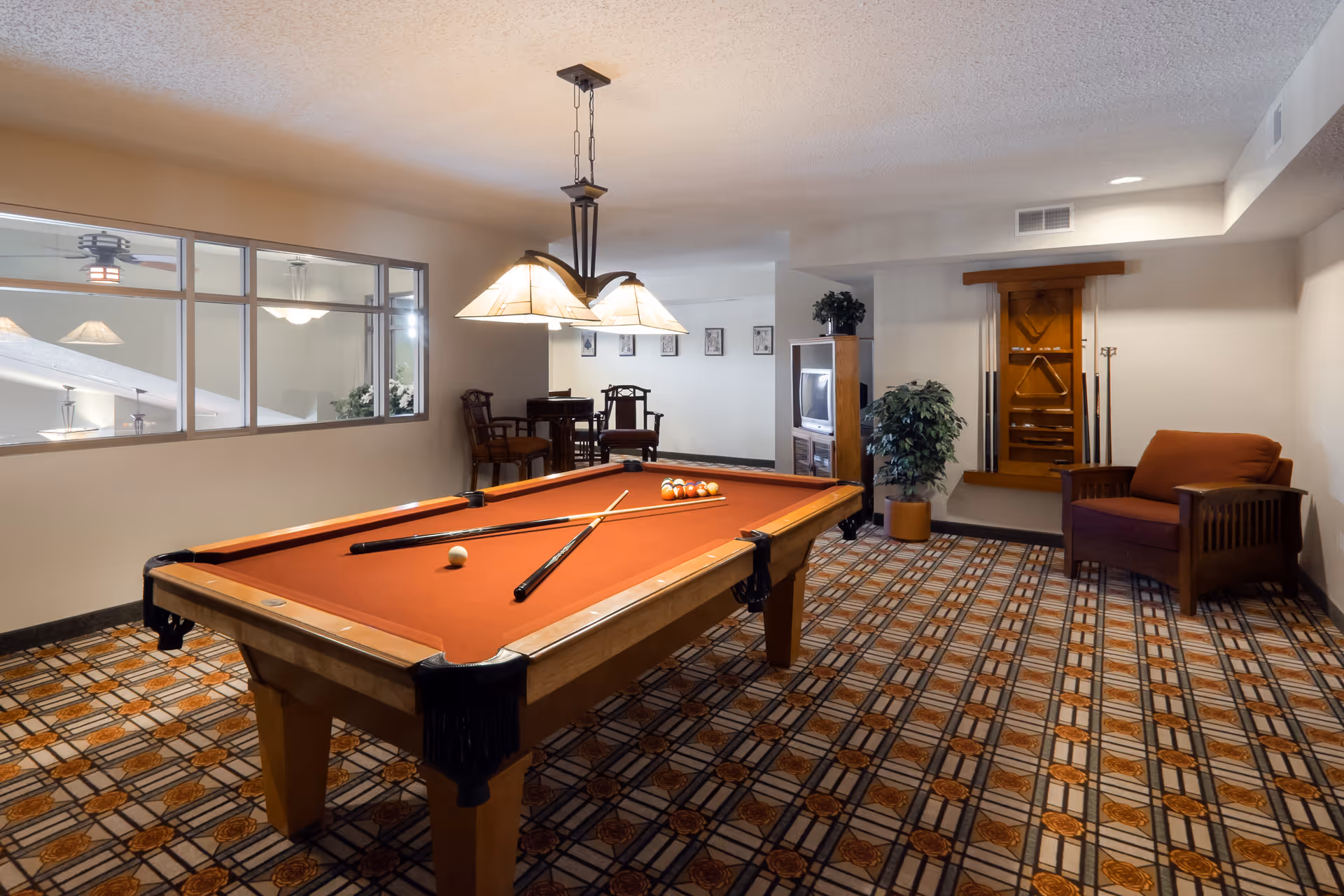 A recreational room with a pool table in the center, featuring a red felt surface with pool cues and balls arranged on it. The room has patterned carpet flooring, a hanging light fixture above the pool table, a wooden chair with cushions, a small table with chairs in the background, a TV on a wooden stand, a potted plant, and a wall-mounted rack holding additional pool cues.