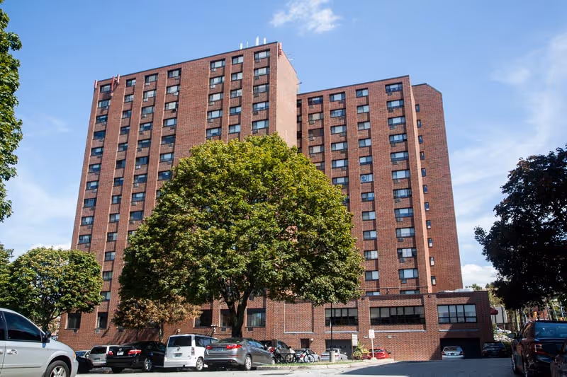 Exterior view of a tall brick senior living facility building with multiple windows, surrounded by trees and parked cars under a blue sky with some clouds.