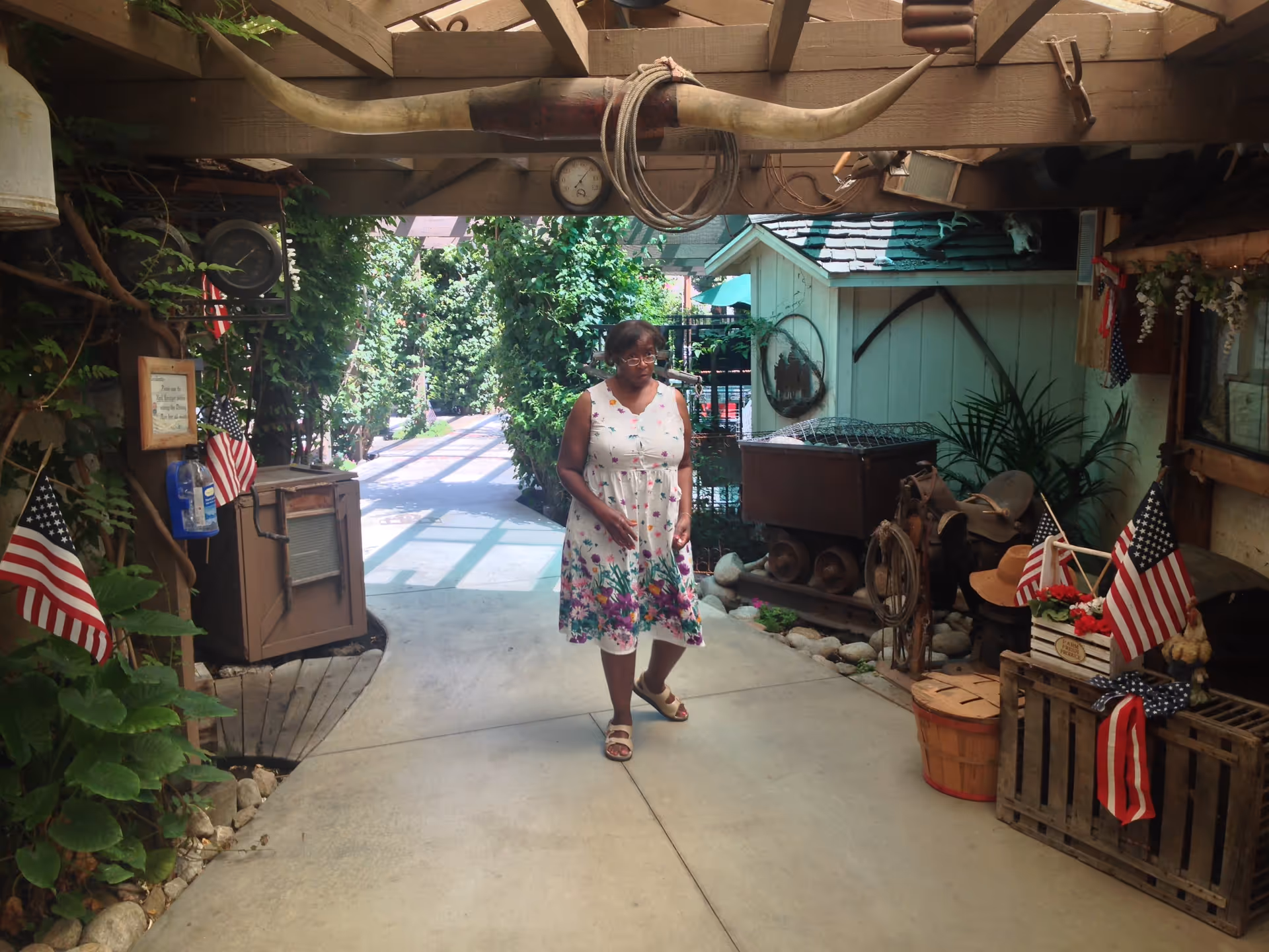 A woman in a floral dress walking through a covered outdoor walkway decorated with American flags and rustic garden decor.
