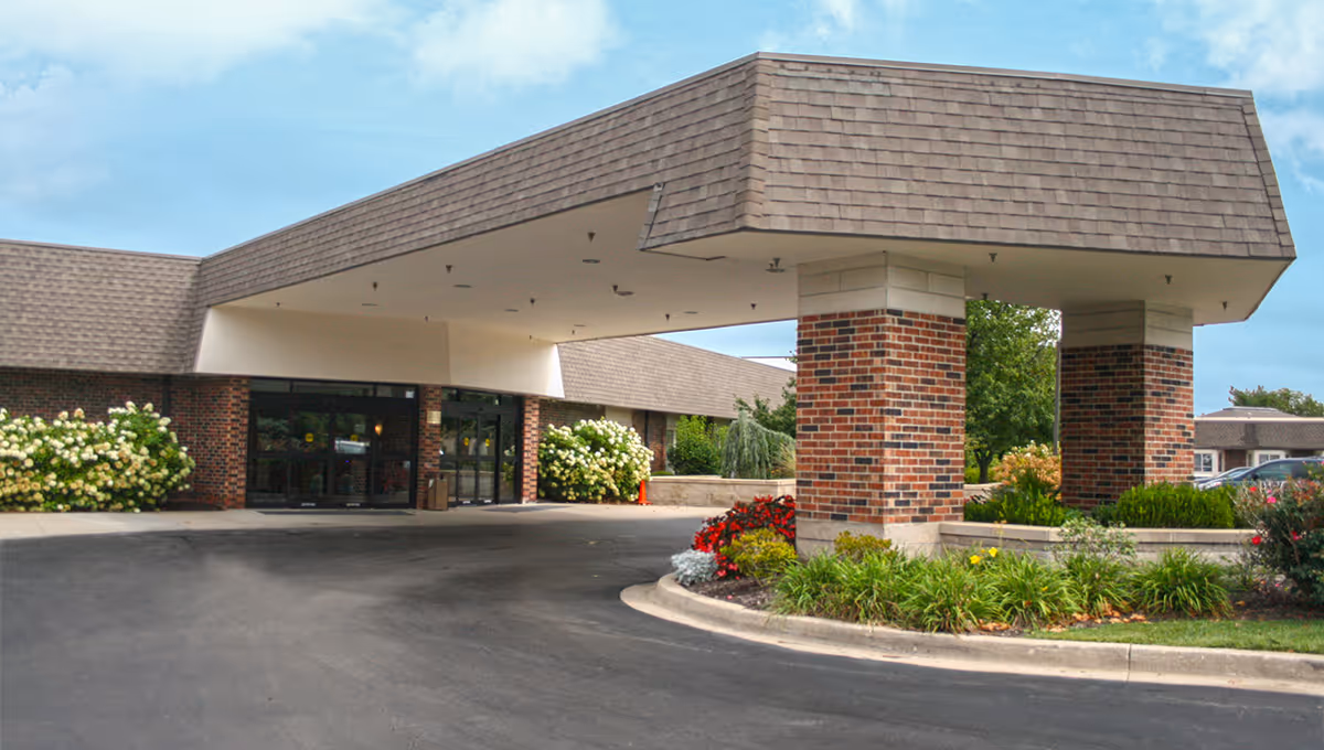 Entrance of Foxwood Springs Senior Living facility showing a covered drop-off area supported by brick columns, with landscaped flower beds and shrubs around the driveway and building.