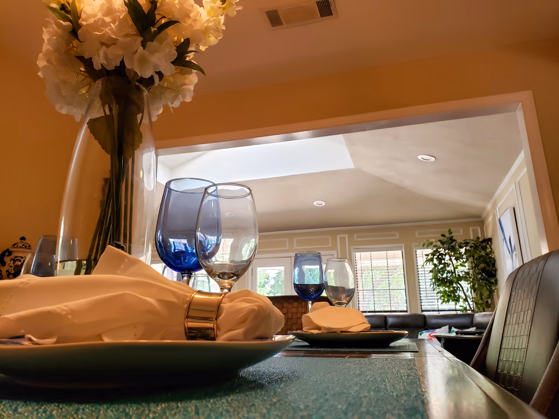 Close-up view of a dining table set with white napkins in silver napkin rings, blue and clear wine glasses, and a large vase of white flowers. In the background, there is a bright living room with large windows, a black leather sofa, and a green potted plant.