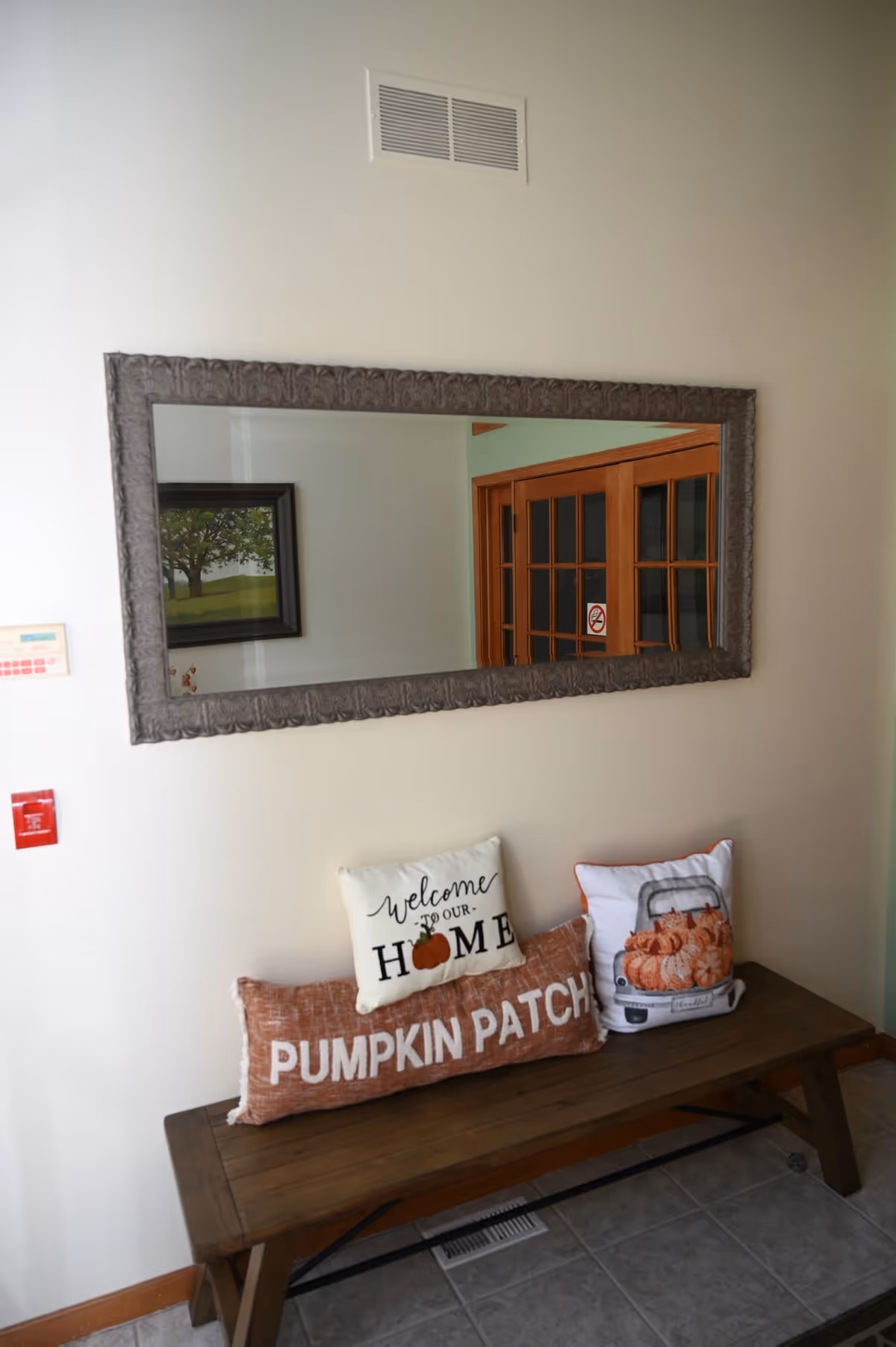 A wooden bench with three decorative pillows placed on it, one reading 'Welcome to our Home' with a pumpkin graphic, another long pillow reading 'Pumpkin Patch', and a third pillow with an image of pumpkins in a truck bed. Above the bench is a large rectangular mirror with a decorative frame reflecting a painting of a tree and wooden framed glass doors.