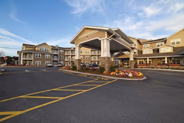 Exterior view of Fairview Estates Gracious Retirement Living facility showing a large covered entrance with stone pillars, surrounded by a multi-story building with beige siding and white trim under a partly cloudy sky.